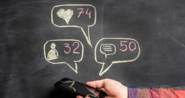 A person holding a smartphone. In chalk on the wall behind it are speech bubbles with likes, comments, and followers symbols