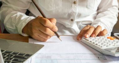 A person at a desk covered in paper using a calculator and a pencil